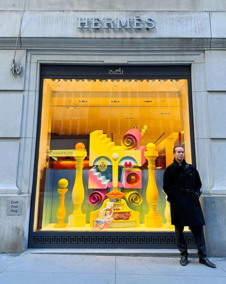 A man in a black coat stands in front of a colorful Hermes window display