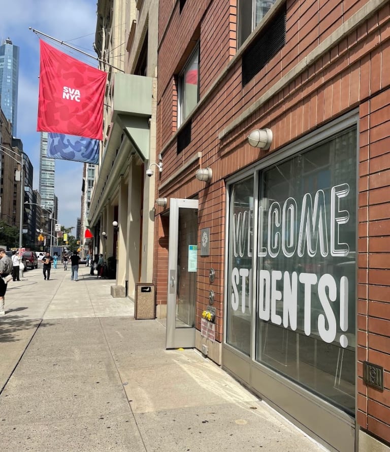 The facade of the East 23rd Street SVA location. A window features a decal that reads "Welcome Students" and two flags—one blue and one red—that say "SVA NYC" hang from the building.