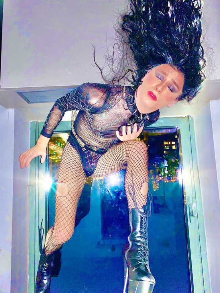 A person in drag stands, leaning forward on their left knee and throwing their long hair in the air. They're wearing metallic platform boots, fishnets and mesh, and facing the camera with their eyes closed.