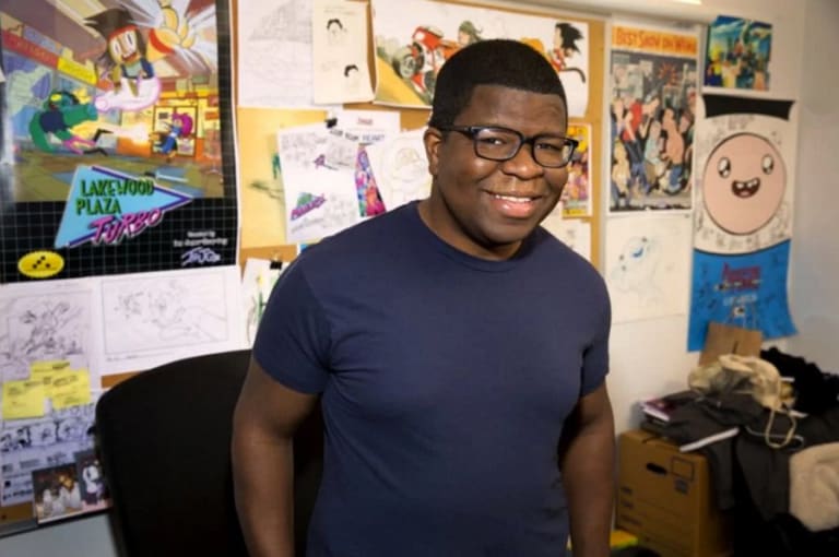A young man in a black t-shirt and black plastic rimmed glasses id's standing in front of a bulletin board full of cartoon images smiling.
