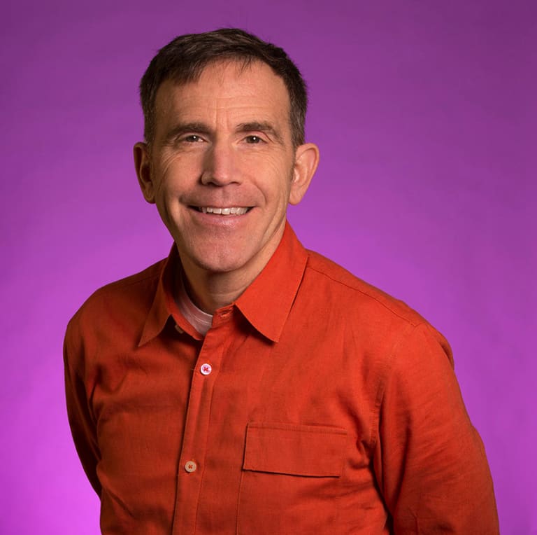A white middle-aged man wearing an orange shirt on a purple background.