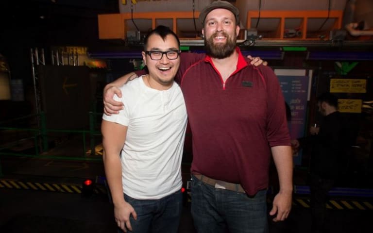 A man with glasses and a white shirt and a man with a hat and beard with their arms around each other's shoulder.