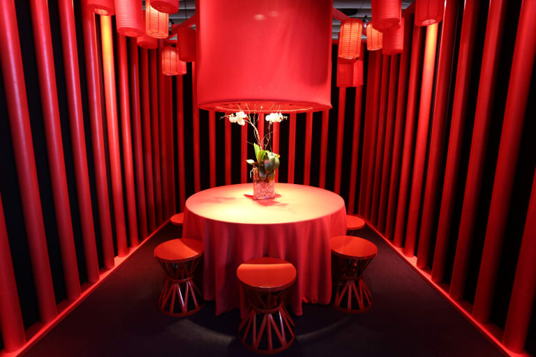 Empty room with walls covered by red drapes. a round red table in the center of the room with a floral arrangement on top and red paper lanterns hanging above it.