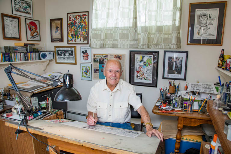 A man sits in front of a drawing board. The room is crowded with works of art and art supplies.