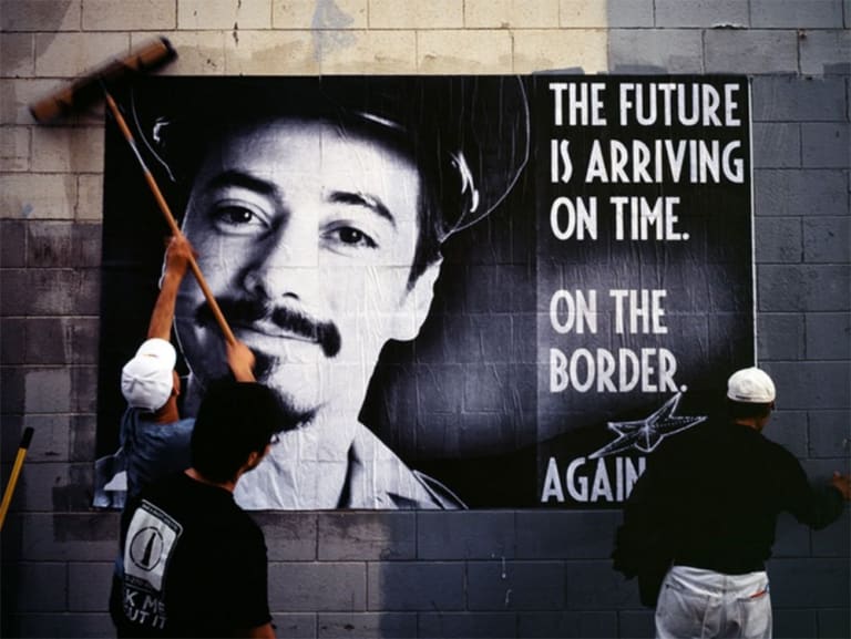 Two men are pasting up a poster while a third watches. The poster reads 