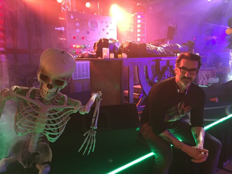 A color photo of a man sitting next to a prop skeleton.
