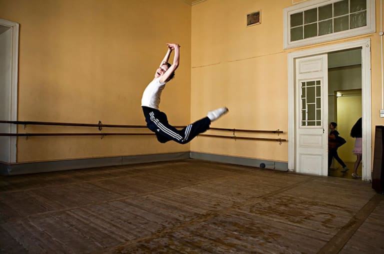 <p "="">A color photograph of a boy smiling and leaping in a dance studio.
