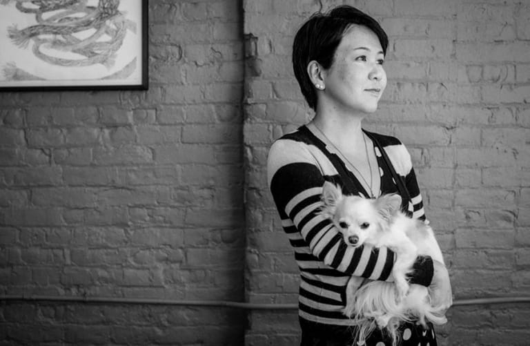 Woman wearing striped shirt holding a dog in front of a brick wall with art hanging on it.