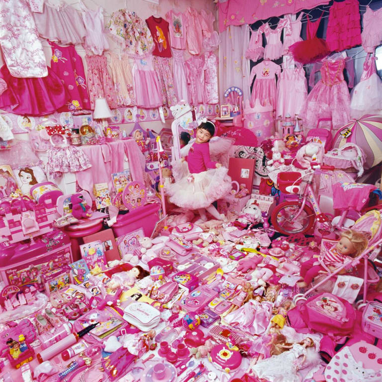 A young girl stands in a room covered with pink clothes and pink toys