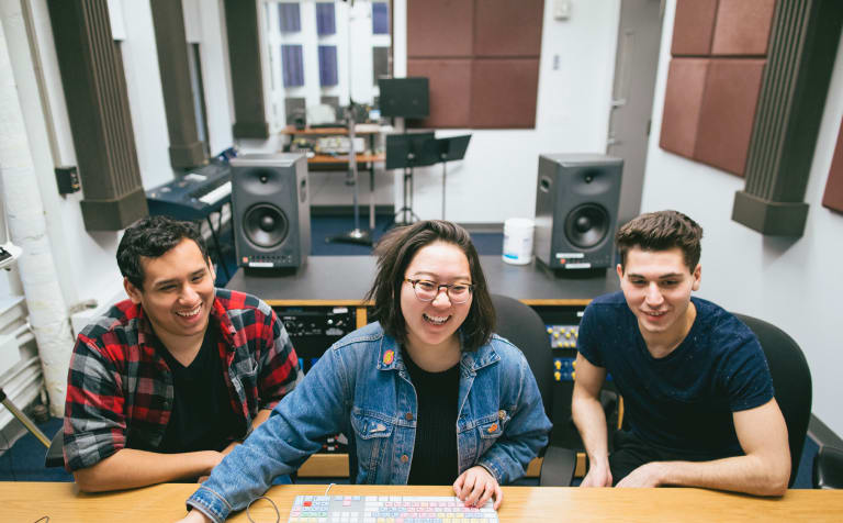 Two young men are sitting on either side of a young Asian woman who is wearing a jean jacket. They are all at a desk or table with a computer keyboard in front of the woman. The room they are in is an office type set-up with musical keyboard, speakers and stands for music toward the back.