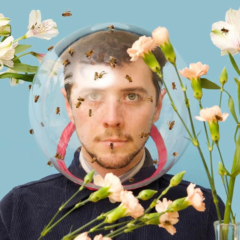 <p "="">A man surrounded by bees and flowers wears a protective glass helmet.
