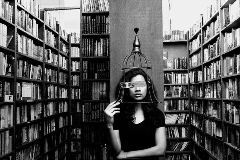 A woman in a library covered in books. She has a cage over her face. Her eyes are offset from her face and replaced with words.