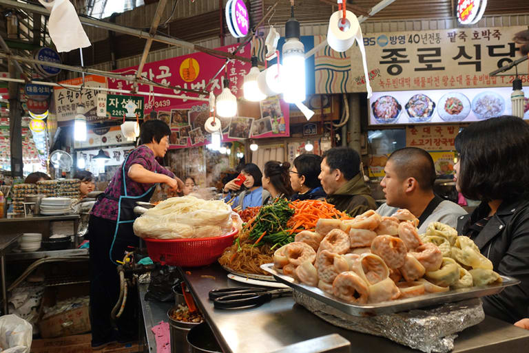 An image of seven people watching a woman cook behind a counter in South Korea.
