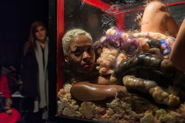A black young woman naked inside a glass cube, sitting in a wet bed of foam and covered in large pieces of synthetic foam.