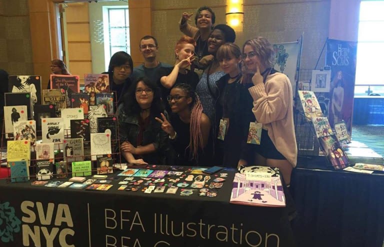 <p "="">A color photo of a group of students gathered behind a table featuring comics and other merchandise.
