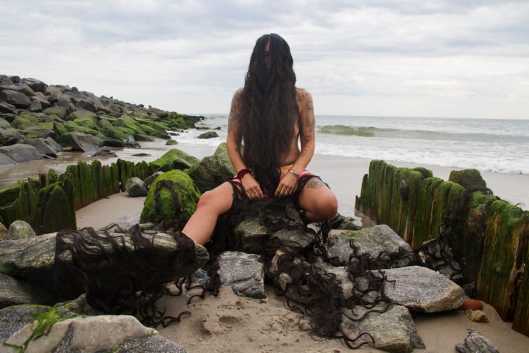 <p "="">a person sits among rocks on a beach, long hair covering their face and chest
