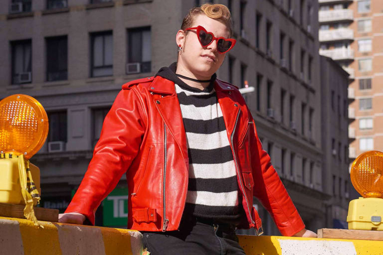 A young man wearing a red leather jacket, black-and-white striped shirt and heart-shaped sunglasses.
