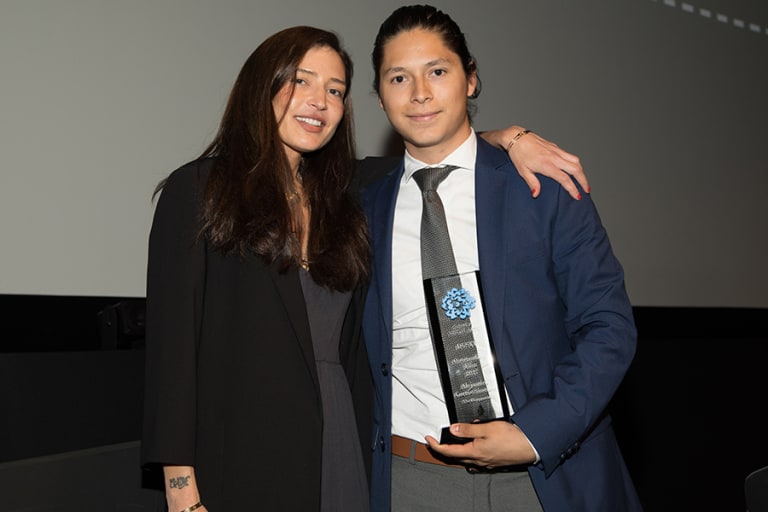 A woman and man standing together; the woman has her arm around the mans shoulders. The man is holding an award.