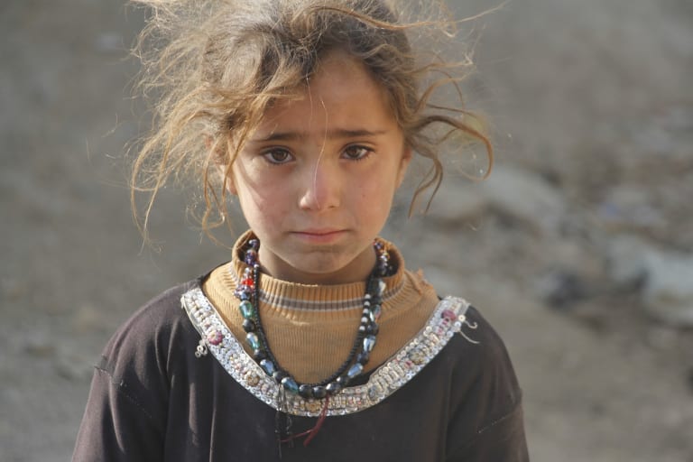A medium portrait of a young Afghan girl with messy hair in partially torn clothing wearing a multi-colored beaded necklace who stares directly into the camera.