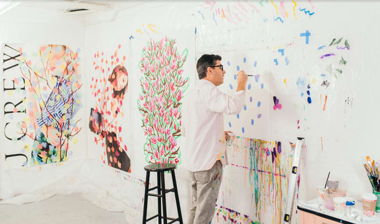 A man painting in a room. The paintings are of J. Crew, two include women, and some have flowers.