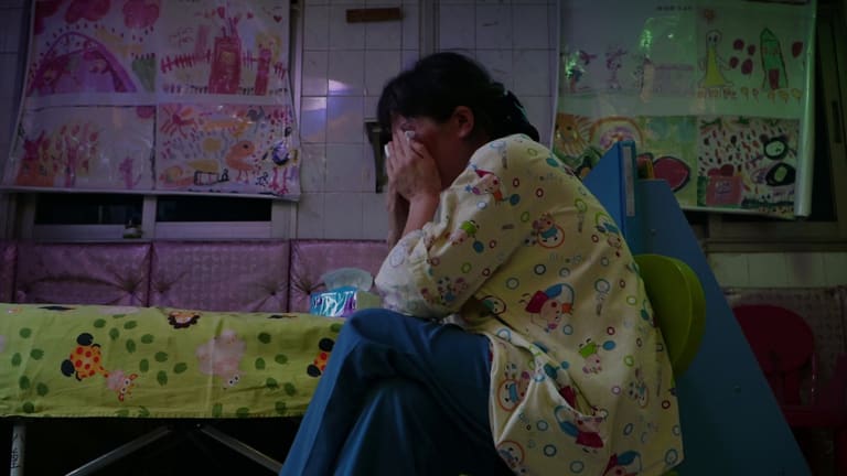  In a tiled room with colorful children’s drawings covering the closed windows, a woman presses her hands against her face, crying into a tissue. 
