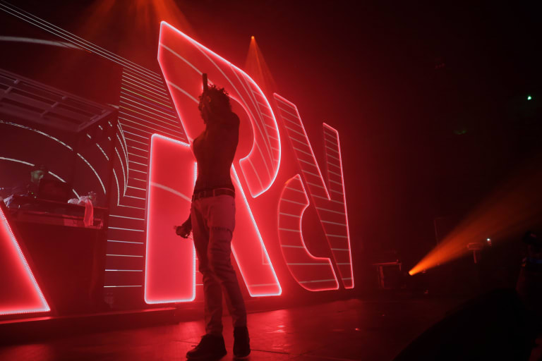 <p "="">A photograph of a performer onstage, accompanied by a DJ, with a large, illuminated logo of Spotify's RapCaviar playlist in the background.
