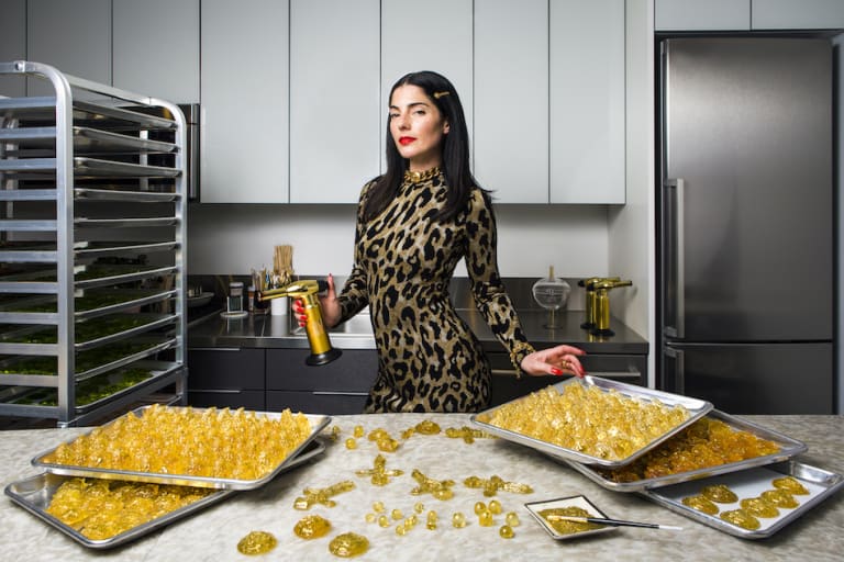 <p "="">A woman in a leopard-print dress stands in a sleek modern kitchen with a gold-plated torch in her hand. On the counter in front of her are several trays of gold-colored candy.
