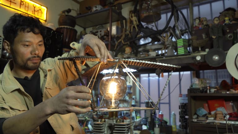 A man builds a sculptural lamp out of leftover motorbike parts in his cluttered studio.