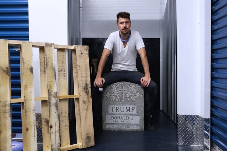 A person sitting on a fake tombstone for Donald J. Trump.