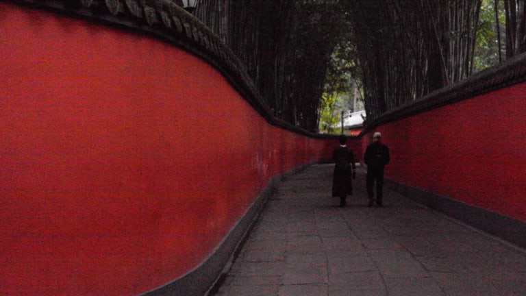 A long winding alley with red walls; in the distance, a couple walks through the pathway. 