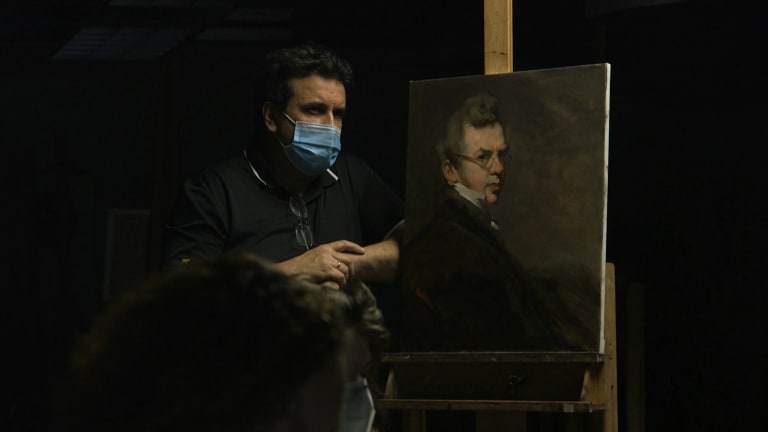 In a dark room, the light highlights a man in a medical face mask with his arm pressing against a classical style portrait painting of a man.