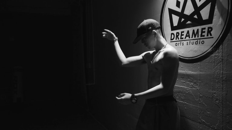 A high contrast B&W image of a young man making a graceful dancer’s pose with his arms. He is shirtless and stands in front of a round sign reading Dreamer Art Studio.