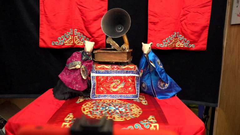 Two mouse-shaped puppets wearing traditional Chinese robes, one purple, one blue, rest against a model gramophone. They are on a stage draped with embroidered red silk.