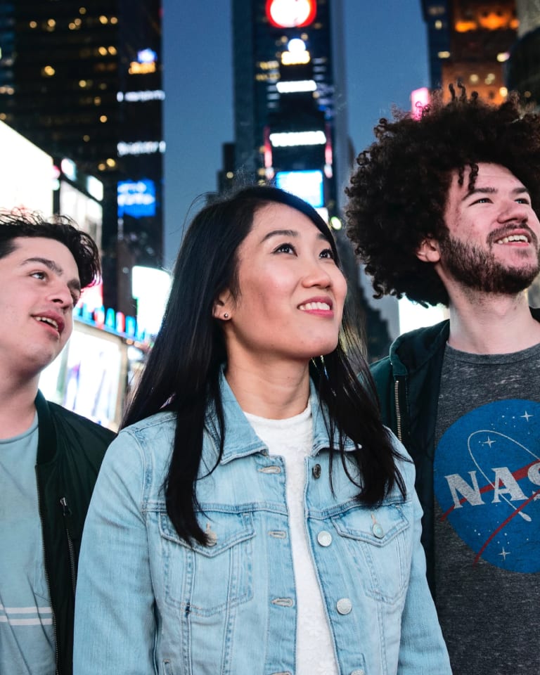 Three people (from left to right; a young white man with short brown hair and a dark green bomber jacker, an east Asian woman with long black hair and a denim jacket, and a taller white man with medium length very curly hair and a beard wearing a grey NASA shirt and hoodie) stand in the middle of Times Square