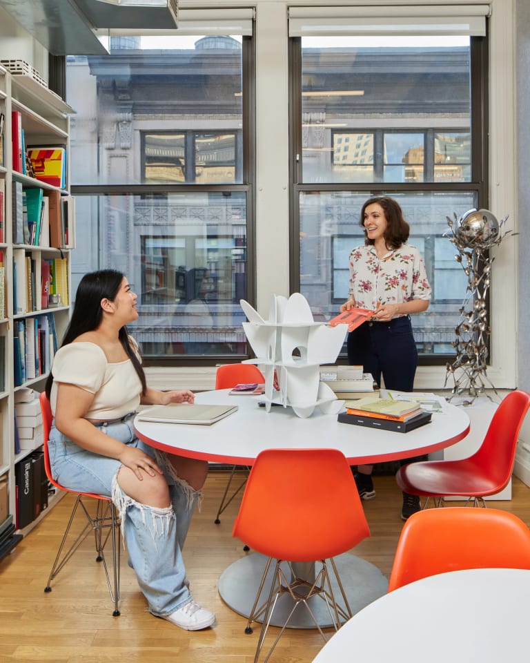 Two people talking in a library; The person on the left is sitting at a table while the person on the right is standing by the window.