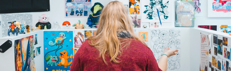 A person with long blonde hair, wearing a red sweater from behind. They're sitting in their studio surrounded by video game, Japanese animation and other kinds of paraphernalia. 