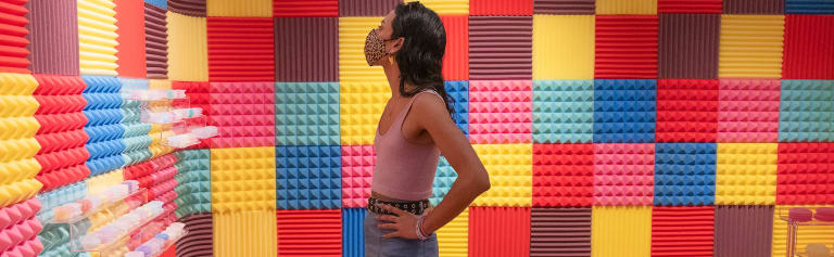 Profile view of a person wearing a pink crop top and a denim skirt with a black studded belt observes the room around them covered in textured tiles in colors of yellow, blue, teal, pink, brown & red
