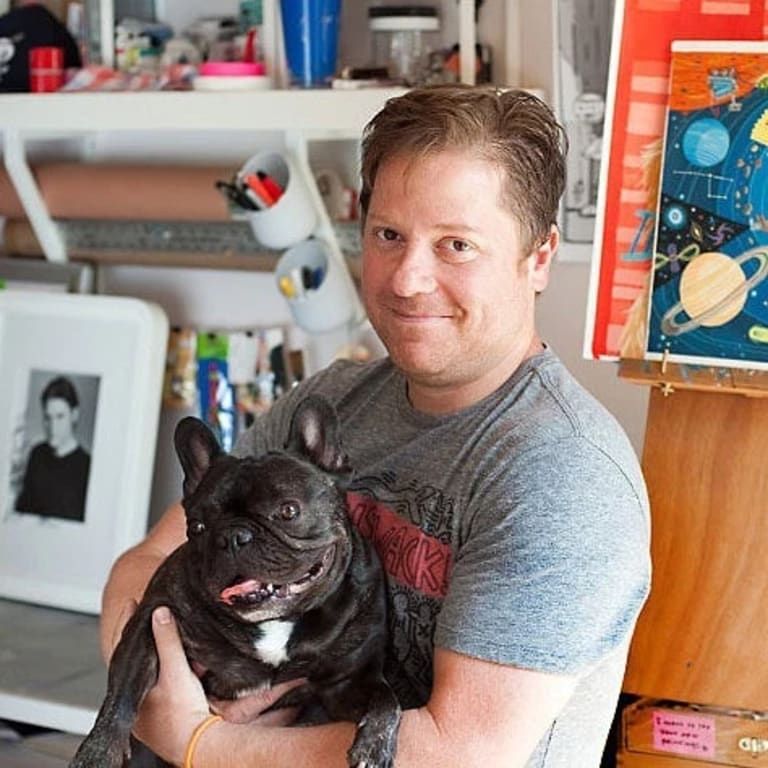 A white man with blonde hair and blue eyes holding a dark brown french bulldog smiling in his studio.