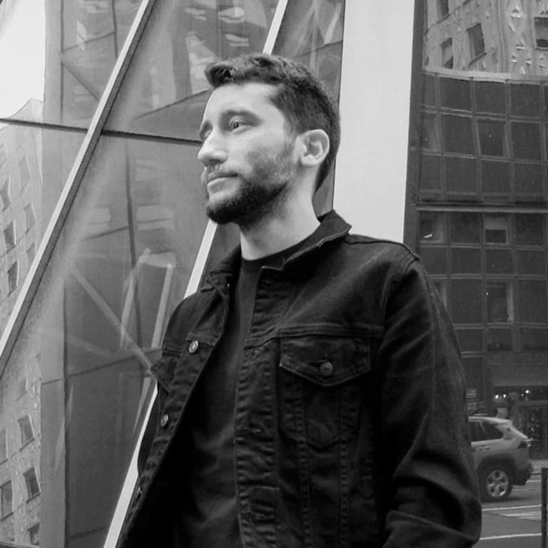 A black and white photograph of a man wearing a dark jean jacket standing in a city setting.