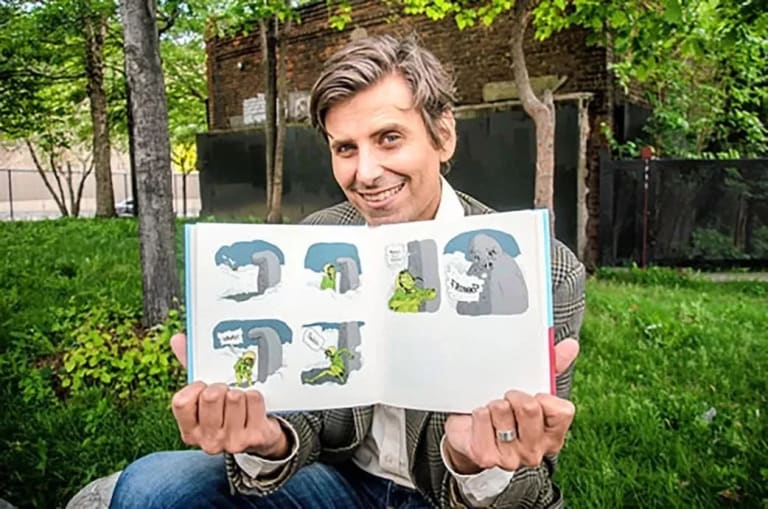 A white man with gray and brown short hair smiles holding a children's book. He is sitting outside in a natural setting with a brick building behind him.