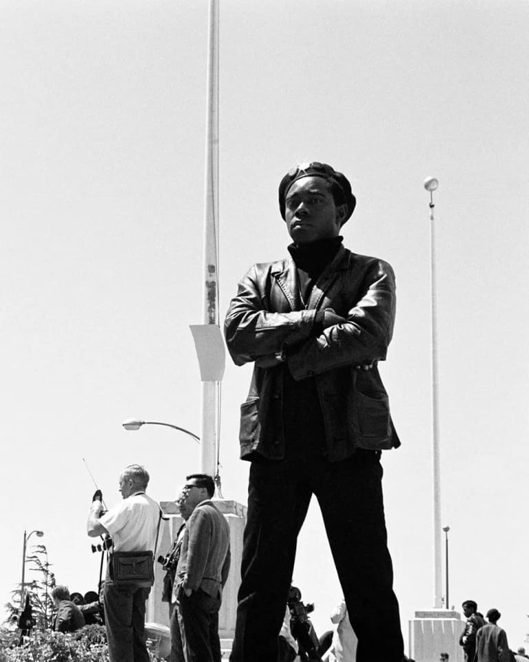 A B&W photograph of three members of the Black Panther Party in Black leather jackets Black berets standing guard outside with a flagpole in the background.