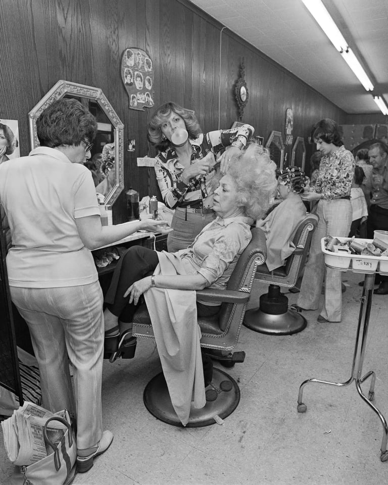 Artist’s mother in a beauty salon getting her hair teased while the beautician blows bubble gum.