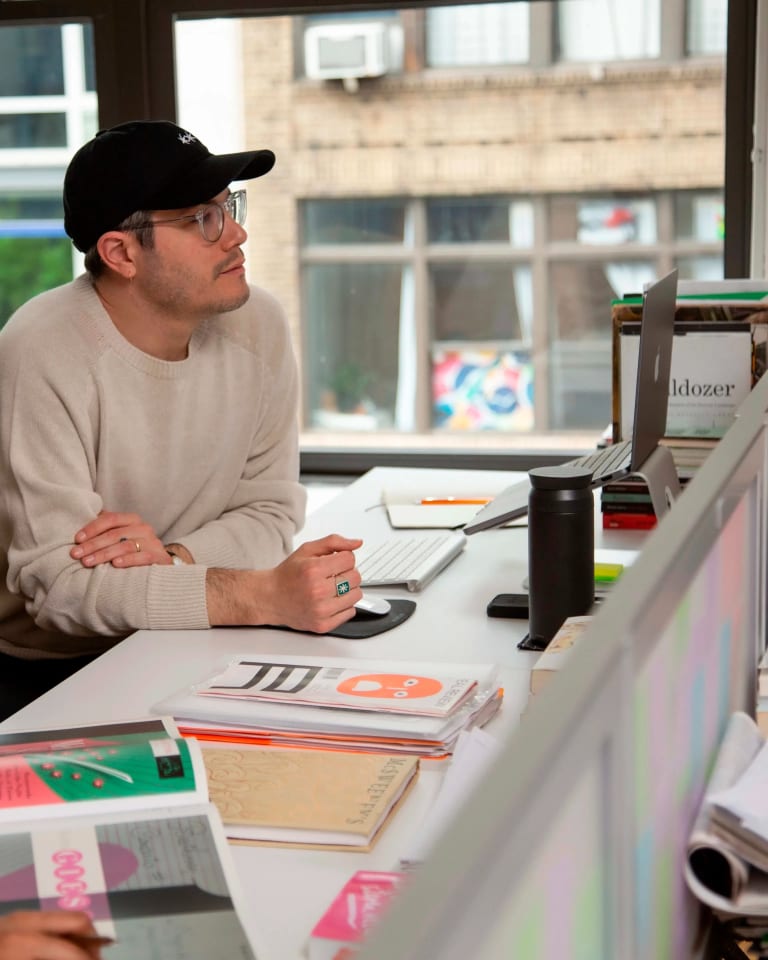 A man in a baseball cap sits at a desk in a bright room next to a woman with long brown hair surrounded by books.