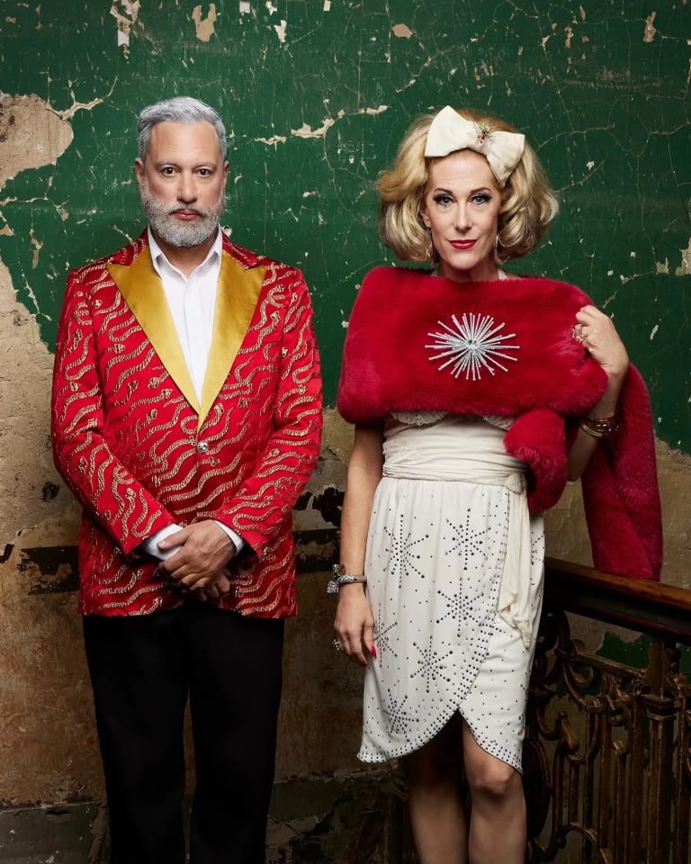 Image of a cis queer gendered man in a red patterned suit with a yellow collar and a them/ they drag performer in a white dress and red fur stole standing in front of a distressed old stucco green wall.Image of a cis queer gendered man in a red patterned suit with a yellow collar and a them/ they drag performer in a white dress and red fur stole standing in front of a distressed old stucco green wall.v