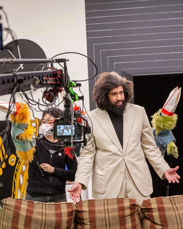 Production still from Jayson Musson's video performance entitled "His History of Art" from 2022. The artist is dressed in a beige suit with a black turtleneck and is performing as "Jay", and standing between two furry puppets, each wearing denim vests, sunglasses and pope's hats. There are production assistants and a camera on an arm in front of him, signaling that this is a behind the scenes shot.