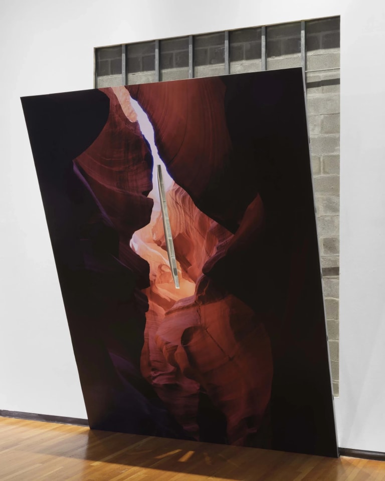 Artwork by Letha Wilson entitled Slit Slot Canyon, 2023. A print of a canyon leans forward off the wall and has a slit in it that looks through to a print of bricks pasted on the wall.Materials are metal studs, drywall, joint compound, digital print on adhesive vinyl, hole in wall. Dimensions are 180" x 110" x 60"