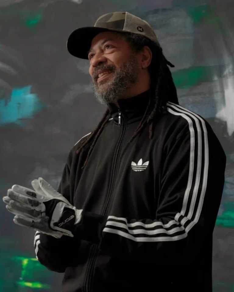 Portrait of a man, Gary Simmons, standing in front of a black wall with white, green and blue paint smudges and starts. The man is wearing a cap, an adidas jacket, and thick gloves.
