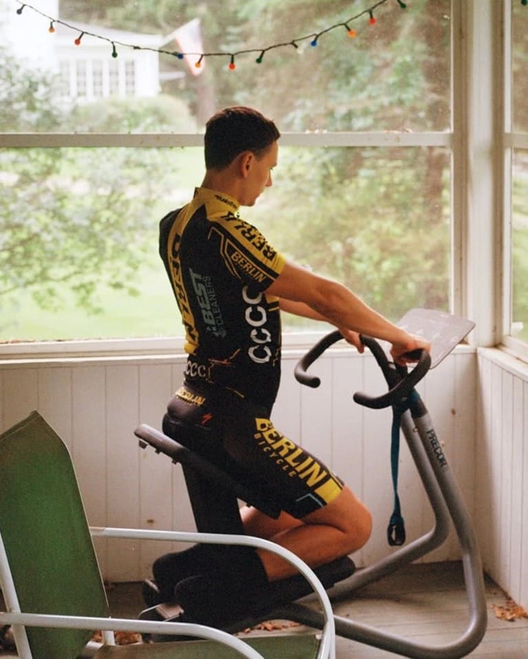 Photograph of a light-skinned man on exercise bike looking at a list of movements while inside an enclosed patio or sunroom.