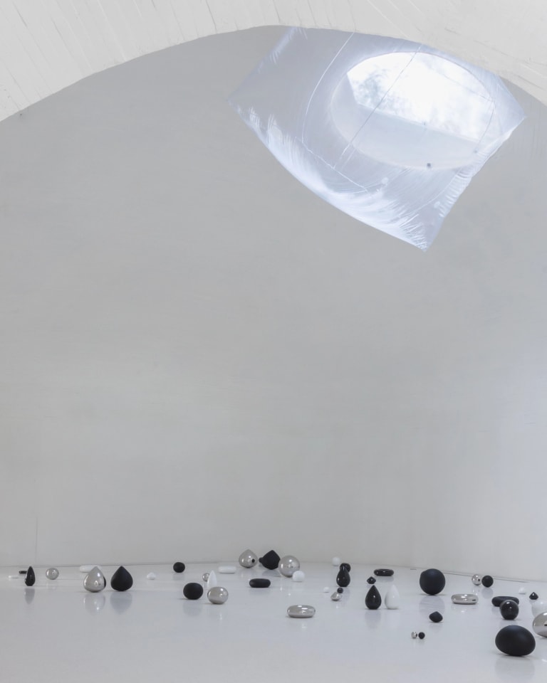 Installation image from the inside of a space with curved architecture, with small black, white, and chrome sculptures clustered all over the floor. Some are orbs, some are tear-drop shaped, and some are flatter like pancakes. They vary in size from an acorn to a basketball.