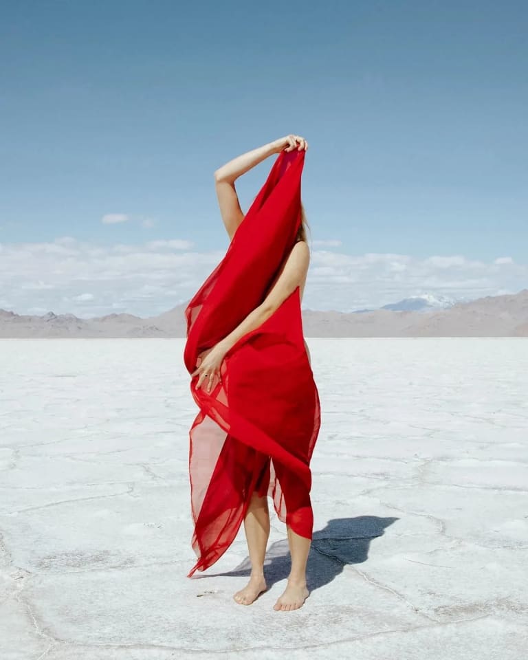 Landscape photograph of a woman holding a red scarf that covers most of her body in the middle of a desert.